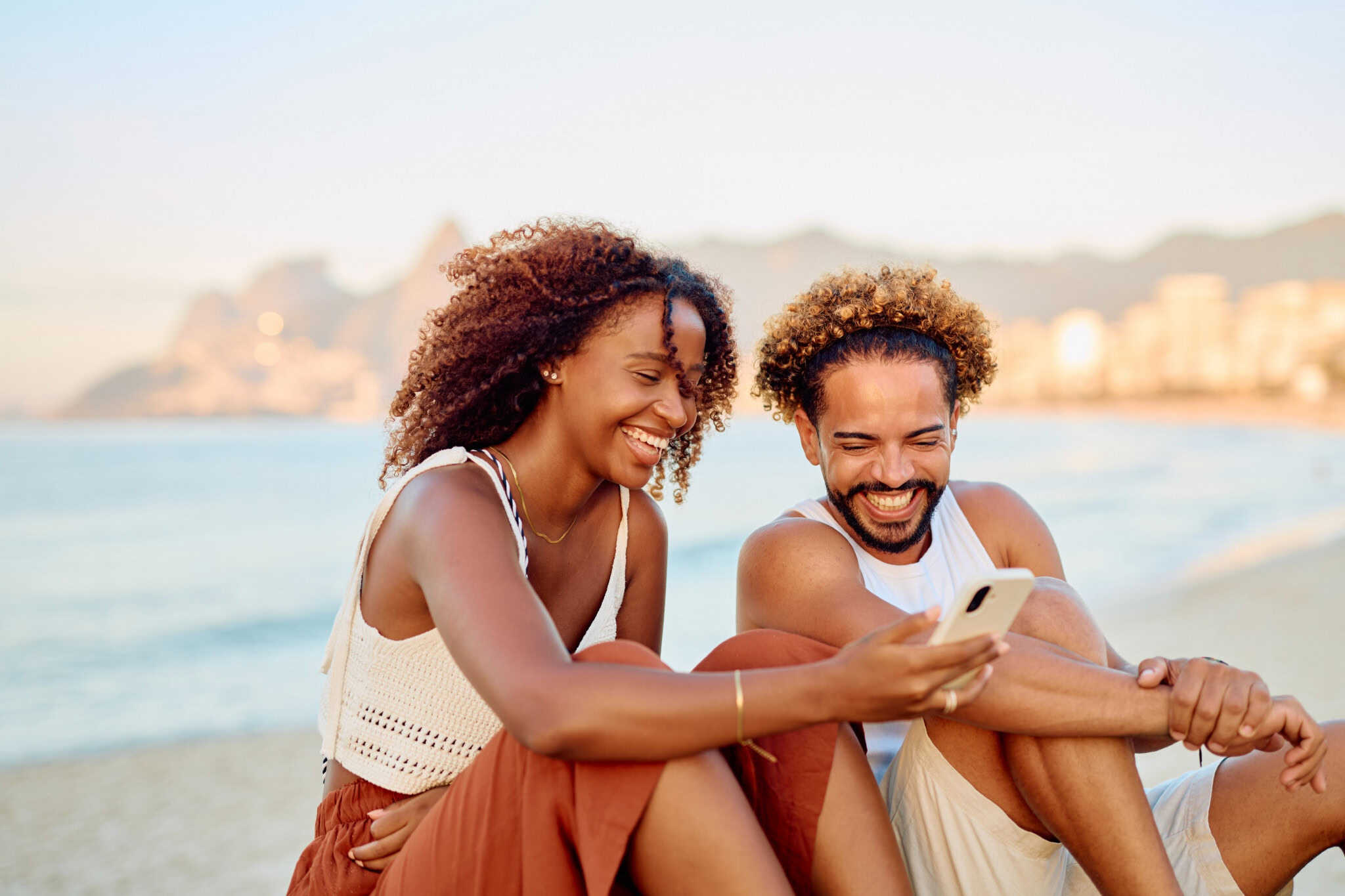 Casal brasileiro em praia olha para o celular enquanto sorriem
