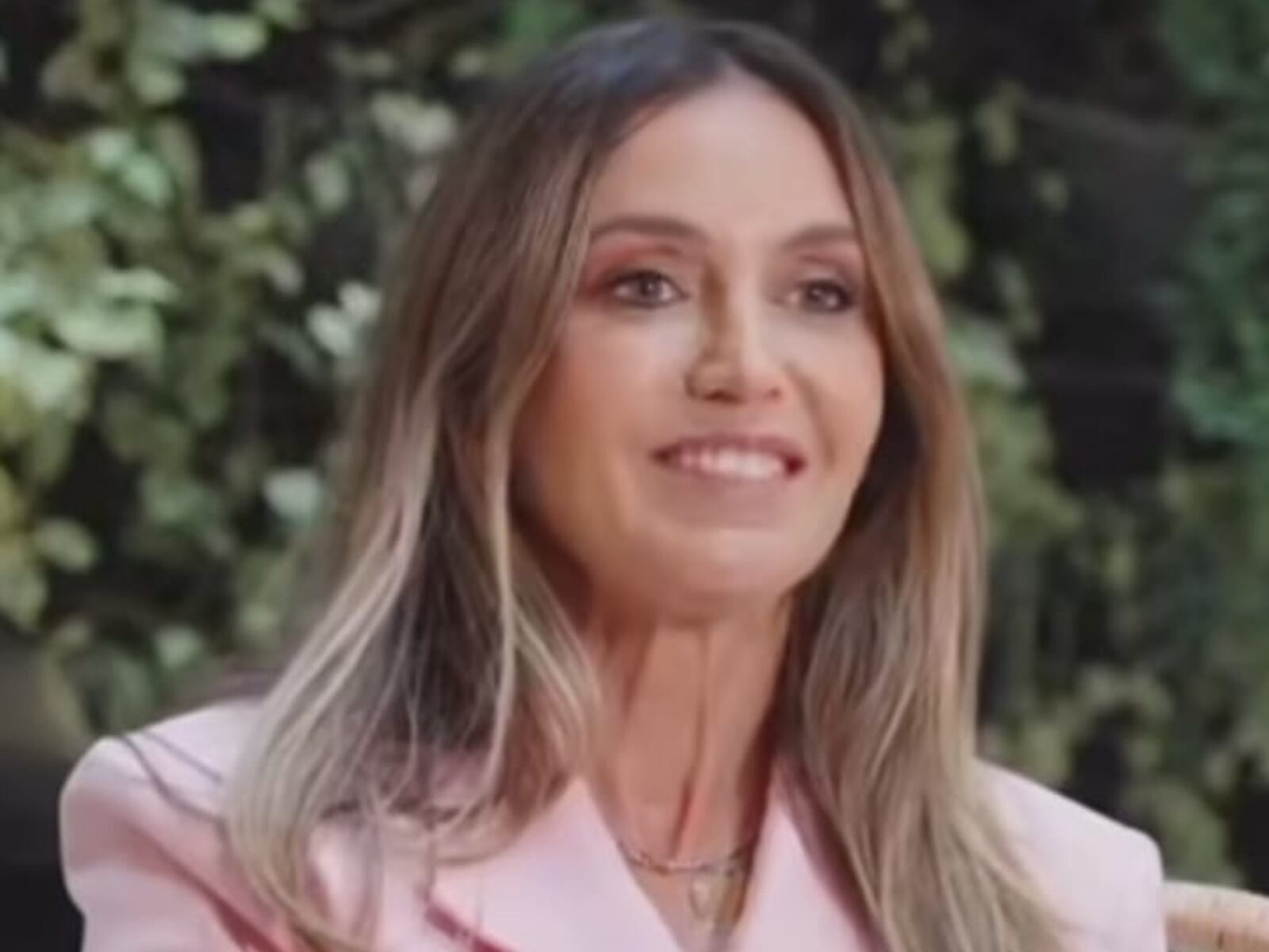 Gabi Medeiros, uma mulher loira e sorridente, veste um blazzer rosa-claro e sorri para a câmera.