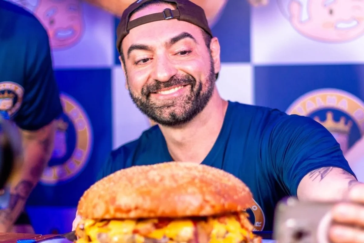 Imagem mostra homem de camisa azul e boné virado para trás em frente a um hamburguer gigante - Metrópoles